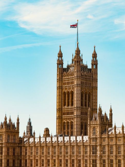 A tall, ornate Gothic-style tower with detailed stonework and multiple spires, topped by a Union Jack flag fluttering in the breeze, set against a partly cloudy sky. The lower portion of the building