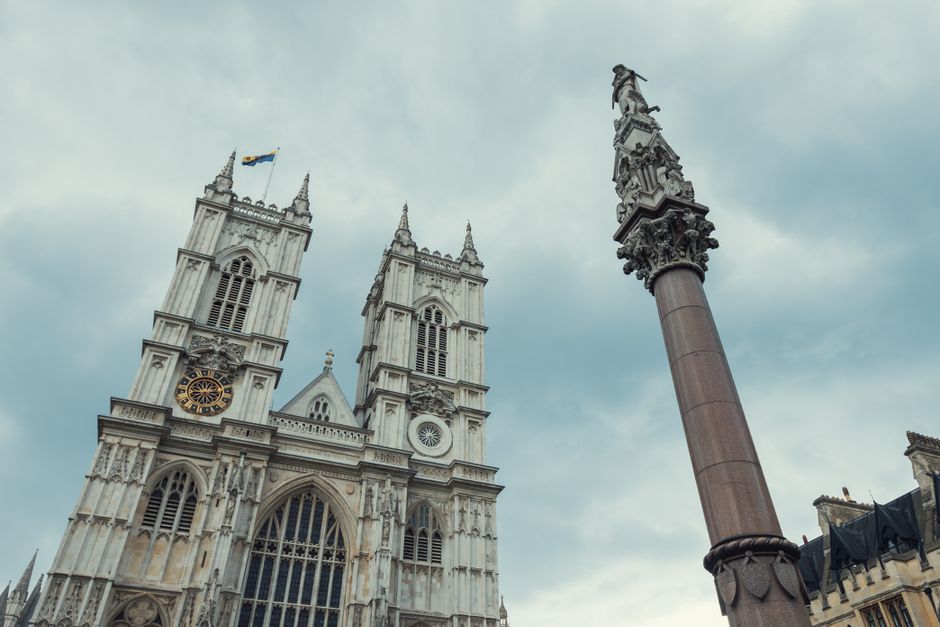 A tall, ornate Gothic-style tower with detailed stonework and multiple spires, topped by a Union Jack flag fluttering in the breeze, set against a partly cloudy sky. The lower portion of the building